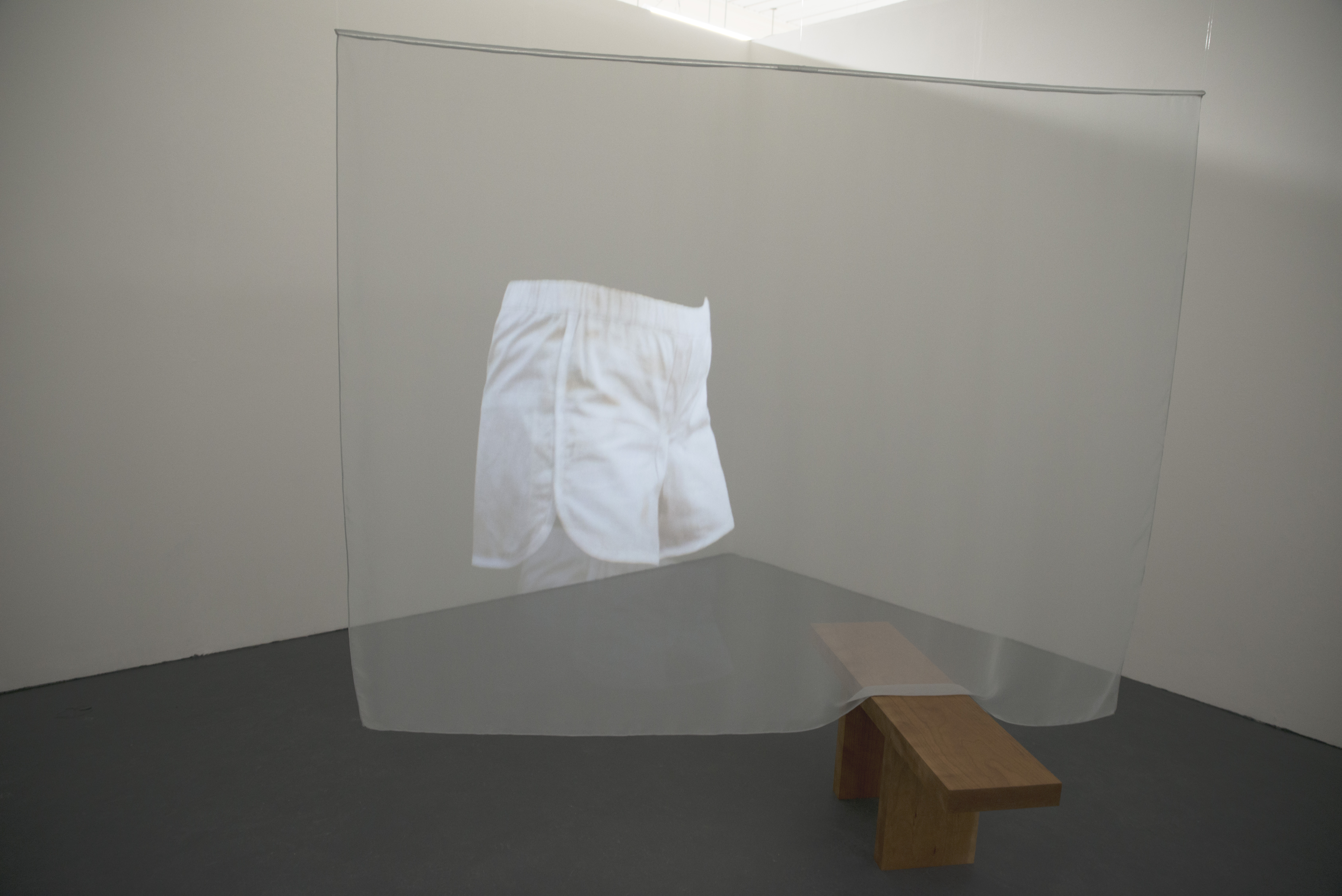 A gallery space filled with a large, human-sized, translucent sheet. This is hanging from the ceiling and one part drapes slightly onto a bench, which perpendicularly intersects the suspended sheet. The large fabric sheet has a white pair of shorts, projected onto it.
